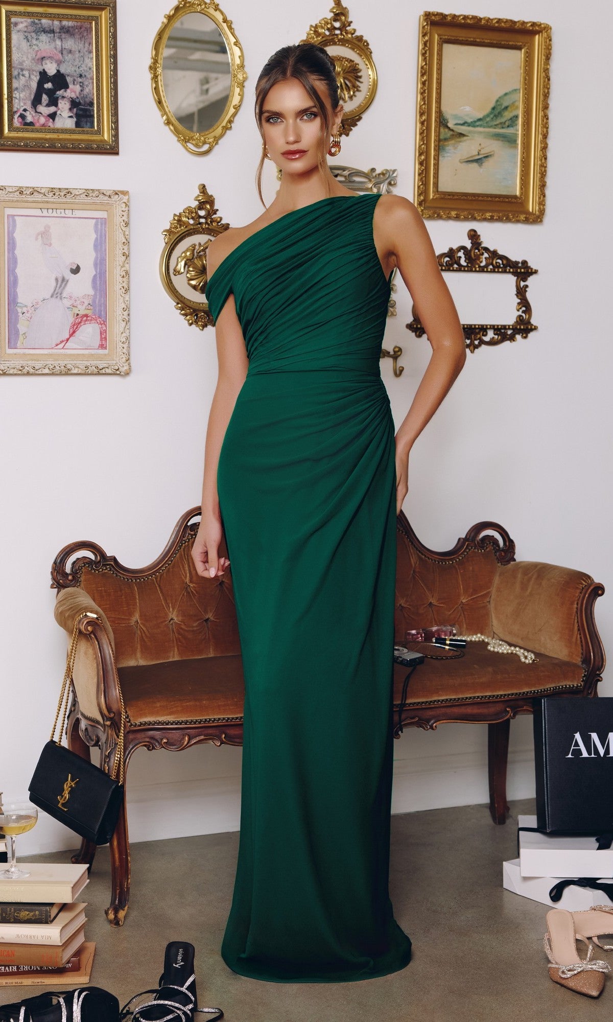 A woman in the Amoris T730 long prom dress in emerald, featuring a one-shoulder ruched chiffon bodice, stands before a vintage loveseat with gold-framed art and mirrors, designer shopping bags scattered nearby.